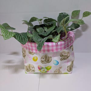 May include: A fabric planter pot with a white background featuring a pattern of bunnies, Easter eggs, and flowers. The top edge is lined with pink and white gingham fabric. The pot contains a green plant with white veined leaves.