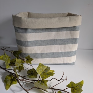 May include: A striped storage basket in light grey and off-white, with a folded-over top edge. The basket has a rectangular shape. A decorative vine with green leaves is in the foreground. The basket is suitable for storage.