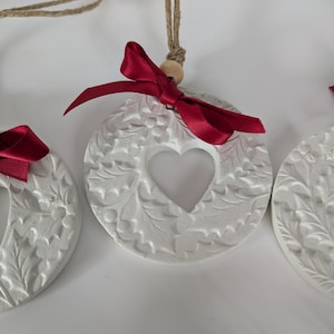 May include: A set of circular white ornaments featuring an embossed holly leaf and berry pattern with a central heart-shaped cutout. Each ornament is adorned with a deep red satin ribbon bow and hangs from a natural jute twine string.