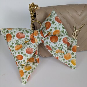 May include: A large fabric bow featuring a white background with a repeating pattern of orange pumpkins and green foliage. The bow is attached to a gold-toned metal ring and is positioned in front of a beige quilted handbag with a gold metal logo.