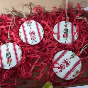 May include: Four circular white ornaments featuring red vertical stripes and illustrations of nutcracker figures. Each ornament has a jute twine loop attached with a small red bead at the top, designed for hanging as decorative holiday accents.