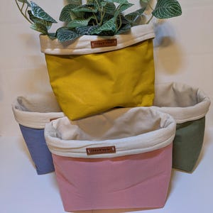 May include: Four fabric storage baskets in yellow, pink, blue, and green, with a white interior and a brown "Handmade" label. The yellow basket holds a green plant with white-veined leaves. The baskets are folded over.