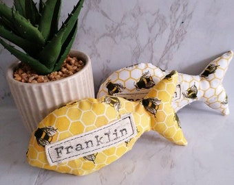 Puede incluir: Dos juguetes de peluche con forma de pez de color amarillo y blanco con un patrón de panal y ilustraciones de abejas. Un pez tiene el nombre "Franklin" bordado.