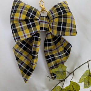 May include: A large fabric bow keyring featuring a yellow, black, and white tartan pattern with thin red and blue accent lines. The bow is attached to a gold-toned metal lobster clasp and ring hardware, providing a secure attachment point.