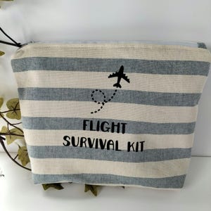 May include: A striped canvas travel pouch in blue and white. The text "FLIGHT SURVIVAL KIT" is printed below a black airplane graphic with a dotted flight path. The pouch has a zip closure.