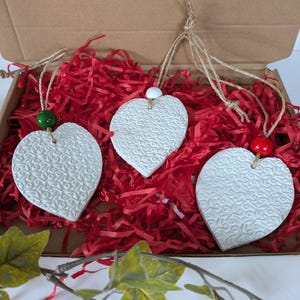 May include: Three white heart-shaped ornaments with a textured floral pattern, each hanging from a jute twine loop. The left heart features a green bead, the centre a white bead, and the right a red bead, all nestled in red shredded paper.