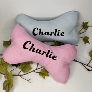 May include: Two bone-shaped dog pillows, one pale blue and one pink, are shown. The name "Charlie" is printed in black on each pillow. Green vines and leaves are arranged around the pillows, creating a decorative display.