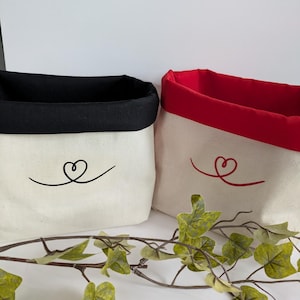 May include: Two canvas storage baskets with a heart design. One has a black rim and heart, the other a red rim and heart. The baskets are off-white and sit on a white surface with a decorative vine.