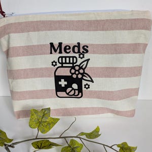 May include: A zipped pouch made of cream and dusty rose striped fabric. The front features a black graphic of a medicine bottle with a cross symbol, pills, and small flowers, with the word "Meds" printed in black text above the bottle graphic.