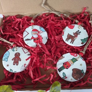 May include: Four circular white ornaments featuring colourful Christmas-themed illustrations including Santa Claus, a reindeer, a gingerbread man, and a nutcracker. Each ornament has a small red bead and a brown twine loop for hanging.