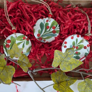 May include: Three circular white ornaments featuring a green mistletoe and red berry pattern. Each ornament is suspended by a brown twine string with a small red bead accent. The ornaments are nestled within a box filled with red paper shreds.