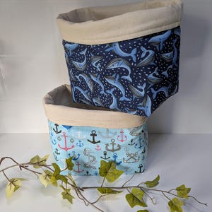May include: Two fabric storage baskets. One basket has a navy blue whale pattern, and the other has a light blue anchor design. Both baskets have a beige rim and are stacked. The baskets are ideal for storage.