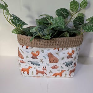 May include: A fabric plant pot with a brown waffle-textured rim and a white body. The body is decorated with woodland animal illustrations, including foxes, squirrels, and bears. Green leafy plants are inside the pot.