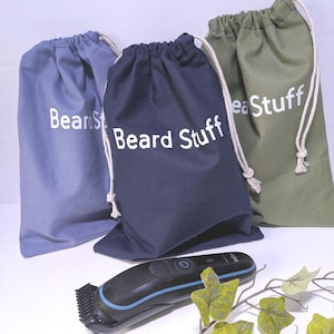 May include: Three drawstring bags in blue, navy, and olive green, each with "Beard Stuff" printed in white. A black and blue beard trimmer sits in front of the bags, with a sprig of artificial leaves.