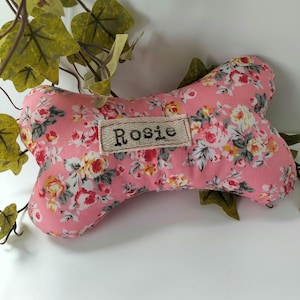 May include: A pink dog bone-shaped toy with a floral pattern of red, yellow, and white flowers. The toy has a small rectangular patch with the name "Rosie" embroidered on it. The background includes green leaves.