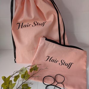 May include: Two pink fabric bags, a drawstring bag and a zippered pouch, both with the text "Hair Stuff" in black script. The drawstring bag has black drawstrings. Several black hair ties and a leafy branch are also present.