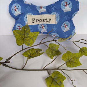 May include: A blue bone-shaped fabric toy featuring a repeating pattern of snowmen inside snow globes and white snowflakes. A rectangular beige fabric label is stitched to the centre with the word Frosty printed in black typewriter font.