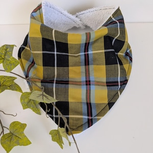 May include: A yellow, black, and blue tartan baby bib with a soft, white, plush lining. The bib is triangular and has a stitched edge. The bib is designed to catch spills and drool.