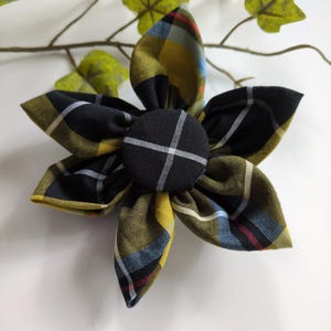 May include: A fabric flower accessory featuring five folded petals made from a plaid textile. The pattern consists of black, mustard yellow, light blue, and thin red lines. A circular fabric-covered button with a black and white grid sits at the centre.