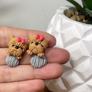 May include: A pair of handmade Yorkshire Terrier dog earrings. Each earring features a brown dog with a gray body and a small red bow. The earrings are displayed on a wooden surface.