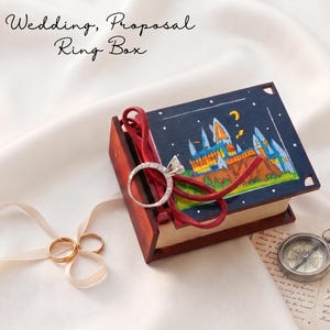 May include: A wooden ring box shaped like a book with a dark blue cover featuring a hand-painted castle illustration under a night sky. The text Wedding, Proposal Ring Box is visible. A silver diamond ring rests on a red ribbon atop the box.