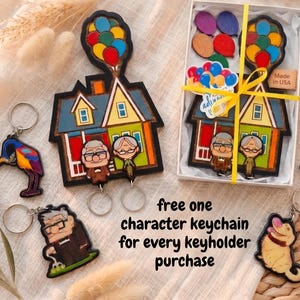May include: A wooden house-shaped key holder with a balloon cluster, featuring two cartoon characters, accompanied by several character keychains and a gift box with a yellow ribbon. Text reads: free one character keychain for every keyholder purchase.