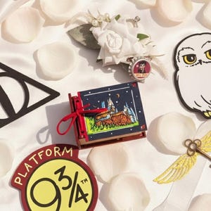 Peut inclure: Divers objets sur le thème d'Harry Potter, dont un symbole des Reliques de la Mort, un panneau de la voie 9 3/4, un petit livre avec une illustration de Poudlard, une découpe de chouette Hedwige et une clé dorée ailée. Des pétales de roses blanches sont éparpillés.