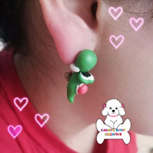 May include: Green and white Yoshi earrings. The earrings are a 3D design of the character Yoshi from the Super Mario video game series. The earrings are attached to an ear. The earrings are green, white, black, and pink.