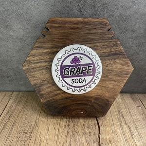 May include: A white button with a purple grape design and the text "GRAPE SODA" on a wooden hexagonal stand.