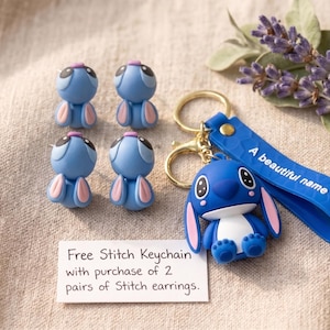 May include: Four blue and pink character stud earrings and a blue character keychain with a gold ring and a blue strap featuring the text A beautiful name. A white card displays the text Free Stitch Keychain with purchase of 2 pairs of Stitch earrings.