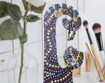 May include: A decorative wooden jewelry stand with a colorful dot pattern in shades of blue, pink, and purple. The stand features multiple levels for hanging necklaces, earrings, and rings. A rose in a glass vase is in the background.