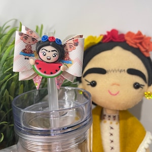 May include: A clear plastic tumbler with a straw, topped with a decorative bow featuring a Frida Kahlo-inspired character holding a watermelon slice. A Frida Kahlo doll is in the background.