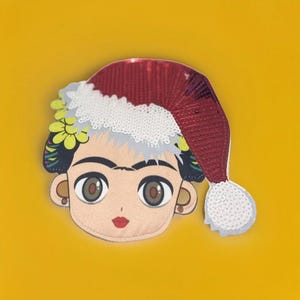 Peut inclure: Un patch en feutre coloré de Frida Kahlo portant un chapeau de Père Noël rouge et blanc à paillettes.