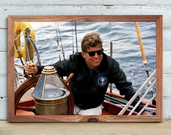 May include: A black and white photo of a man wearing sunglasses and a black jacket, steering a sailboat on the water.