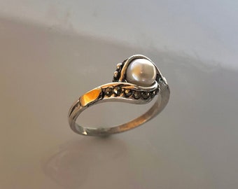 Könnte beinhalten: Ein silberner Ring mit einem weißen Perlmutt, das in einem Wirbelmuster eingefasst ist. Der Ring ist mit kleinen schwarzen Steinen verziert.