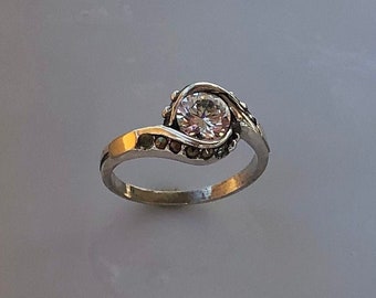 Könnte beinhalten: Ein silberner Ring mit einem klaren runden Stein, der in einem Wirbelmuster eingefasst ist. Der Ring ist mit kleinen schwarzen Steinen verziert.