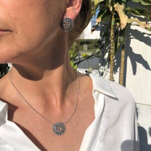 May include: A silver necklace and earrings set with a delicate floral design. The necklace features a round pendant with a detailed floral pattern, while the earrings are small and round with a similar design.