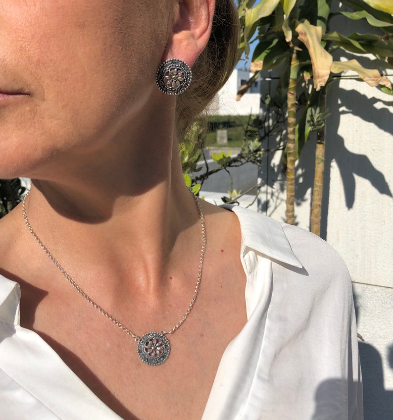 May include: A silver necklace and earrings set with a delicate floral design. The necklace features a round pendant with a detailed floral pattern, while the earrings are small and round with a similar design.