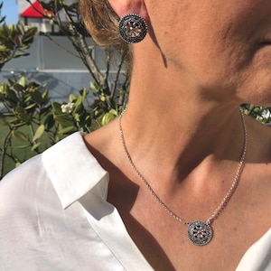 May include: A silver necklace with a round pendant featuring a floral design. The pendant has a black center and silver accents. The necklace is on a silver chain.