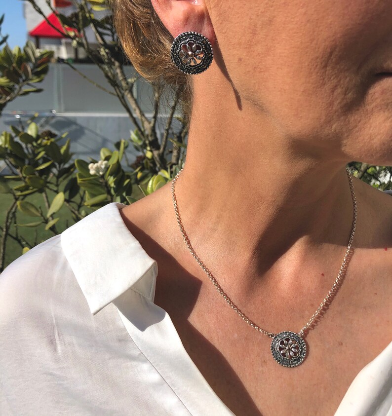 May include: A silver necklace with a round pendant featuring a floral design. The pendant has a black center and silver accents. The necklace is on a silver chain.
