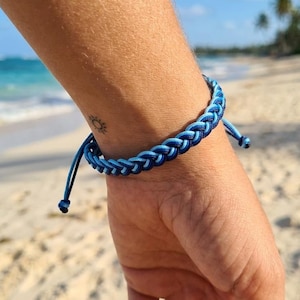 May include: A braided cord bracelet featuring a gradient of blue tones, ranging from light sky blue to deep navy blue. The bracelet is constructed with a woven pattern and includes two thin adjustable cord ends that hang loosely from the main band.