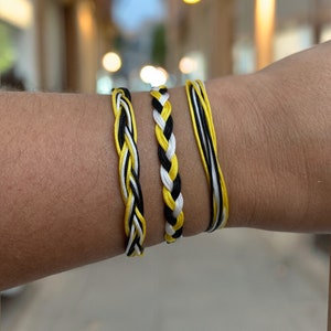 May include: Three braided bracelets in black, white, and yellow are displayed. The bracelets are made of woven threads, with varying patterns of color. The bracelets are worn on a person's wrist, showcasing the jewelry.