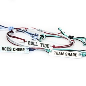 Op de afbeelding: Vier kleurrijke vriendschapsarmbanden met zilveren metalen labels. De labels hebben de volgende tekst: "ROLL TIDE", "TEAM SHADE", "NCCS CHEER".