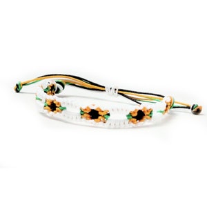 May include: White friendship bracelet with green and orange accents, featuring a repeating pattern of small sunflower designs.