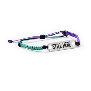 May include: A teal, purple, and black braided bracelet with a silver charm that reads "STILL HERE".