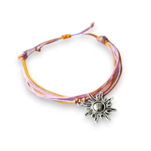 May include: A multi-strand bracelet with a silver sun charm. The bracelet is made of light purple and orange cord.