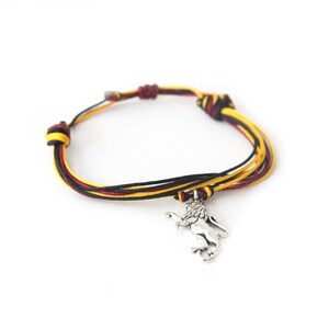 May include: A braided bracelet featuring black, yellow, and maroon cords. A silver-colored lion charm hangs from the bracelet. The bracelet is adjustable and sits against a white background.