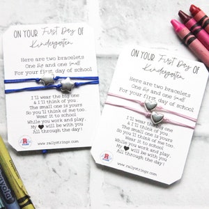May include: Two cards with a blue and pink friendship bracelet on each. The cards have a message for a child's first day of kindergarten. The message reads, "Here are two bracelets, one big and one small, for your first day of school. I'll wear the big one and I'll think of you. The small one is yours so you'll think of me too. Wear it to school while you work and play. My heart will be with you all through the day!"