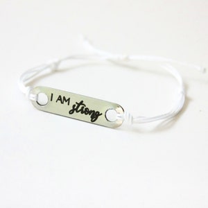 May include: A silver-colored bracelet with the words "I AM strong" in black script. The bracelet features a rectangular metal plate with two holes, connected to a white cord. The cord is adjustable.