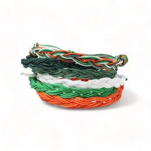 May include: A stack of six braided bracelets in green, white, and orange colors. The bracelets are made of woven string and are stacked on top of each other.